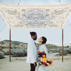 May include: A couple stands beneath a cream-colored canopy decorated with a black and white botanical design. The man wears a white suit jacket and black tie, while the woman holds a bouquet of colorful flowers. String lights are visible in the background.