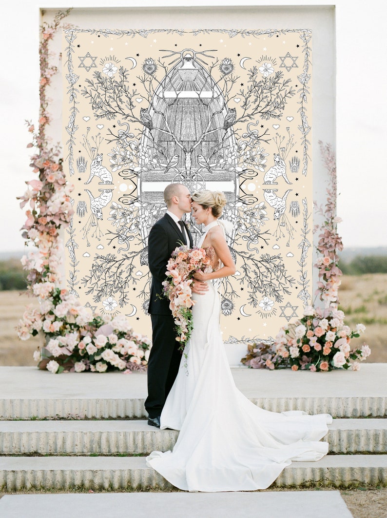 May include: A wedding ceremony scene featuring a couple standing before a decorative backdrop. The backdrop has a cream-colored background with a black and white illustration. The bride wears a white gown, and the groom is in a black suit. Floral arrangements frame the scene.