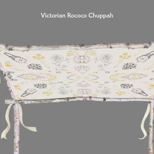 May include: A Victorian Rococo Chuppah with a cream-colored fabric canopy. The canopy is adorned with a repeating pattern of black and yellow floral and abstract designs. The chuppah is supported by birch poles and cream-colored ribbons.