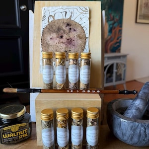 Puede incluir: Una disposición de viales de vidrio con tapas doradas, llenos de hierbas secas y etiquetados. También hay un pincel de madera y un frasco de tinta. Los viales están etiquetados con palabras como "Bienestar" y "Comunidad". Una obra de arte está en el fondo.