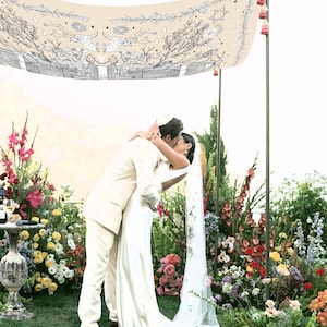 May include: A couple embraces under a cream-colored canopy decorated with black and white illustrations, surrounded by vibrant flowers. The bride wears a white dress and veil, while the groom is in a cream suit. The scene is set outdoors, with a lush floral backdrop.