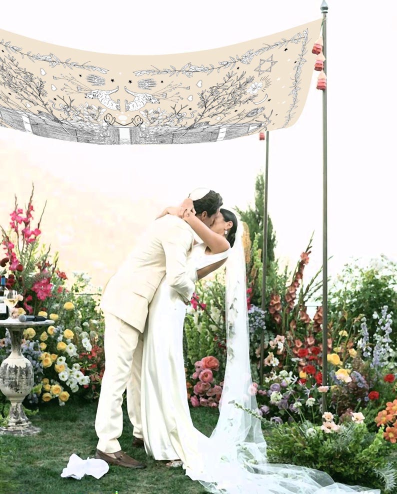 May include: A couple embraces under a cream-colored canopy decorated with black and white illustrations, surrounded by vibrant flowers. The bride wears a white dress and veil, while the groom is in a cream suit. The scene is set outdoors, with a lush floral backdrop.