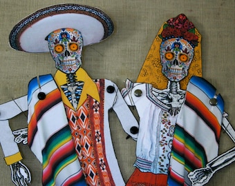 May include: Paper cutouts of a male and female skeleton, dressed in traditional Mexican attire. The male skeleton wears a sombrero and a colorful serape. The female skeleton has a floral headpiece and a veil. The skeletons are decorated with intricate sugar skull designs.