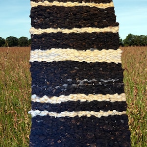 May include: Handwoven rug with alternating bands of black and cream-colored wool. The rug has a rectangular shape and is displayed outdoors against a backdrop of a field and blue sky. The rug has tassels at the bottom.