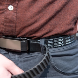 May include: Close-up of a black belt with a metallic buckle. The belt is worn with gray pants and a brown shirt. The belt has the text "CONTITECH CT 588" printed on it.