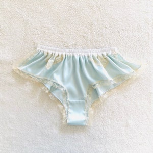 May include: Pale blue satin underwear with a white elasticated waistband and delicate lace trim. The underwear features a ruffled design along the edges and is made of a soft, silky material. The lace trim is a light cream colour.