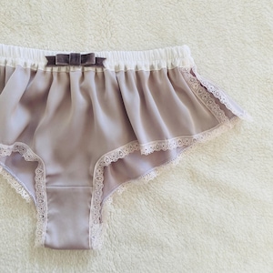 May include: A pair of light purple underwear with a white waistband and lace trim. A small, dark purple bow is centered on the front. The underwear is made of a soft, silky material.