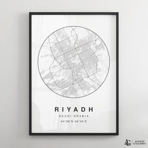 Op de afbeelding: Een zwart-wit ingelijste kaart van Riyad, Saoedi-Arabië. De cirkelvormige stadskaart toont wegen en gebouwen in grijs. De naam van de stad en de coördinaten staan eronder afgedrukt. De lijst is zwart.