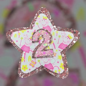 May include: Pink glitter star-shaped applique with a pink number 2 in the centre. The star is decorated with a white background featuring a repeating pattern of pink pigs wearing crowns and holding wands.