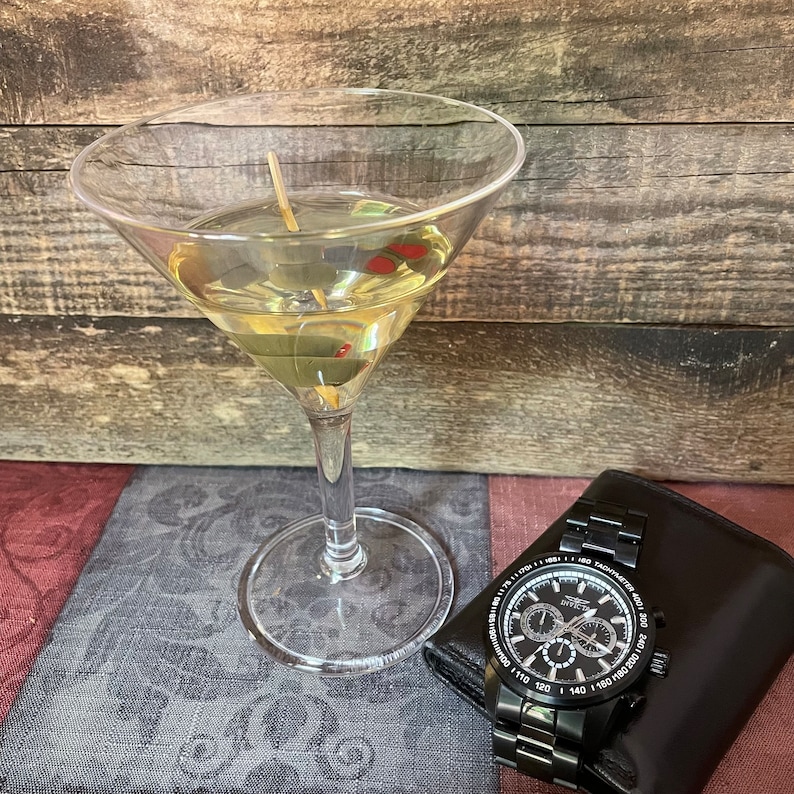Op de afbeelding: Een martini-glas gevuld met een heldere vloeistof en gegarneerd met olijven. Een zwart polshorloge met een zilveren wijzerplaat en een zwarte band ligt op een zwart lederen oppervlak.