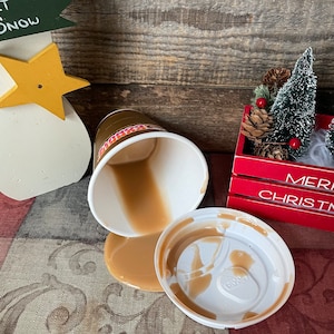 May include: A white coffee cup with a brown lid has spilled its contents on a brown surface. The coffee is a light brown color and has a creamy texture. The cup is sitting on a brown surface with a red and white wooden box in the background.