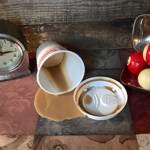 May include: A white coffee cup with a brown lid has spilled coffee on a patterned tablecloth. There are three billiard balls on a red tray in the background.