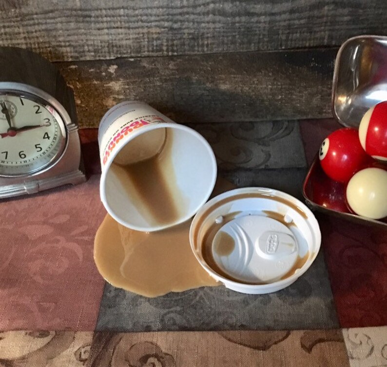 May include: A white coffee cup with a brown lid has spilled coffee on a patterned tablecloth. There are three billiard balls on a red tray in the background.