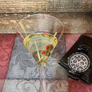 Op de afbeelding: Een martini-glas gevuld met een heldere vloeistof en groene olijven. De olijven hebben rode centra. Het glas staat op een gepatroneerd stofoppervlak. Een zwart horloge met een zwarte wijzerplaat en witte wijzers bevindt zich op de achtergrond.