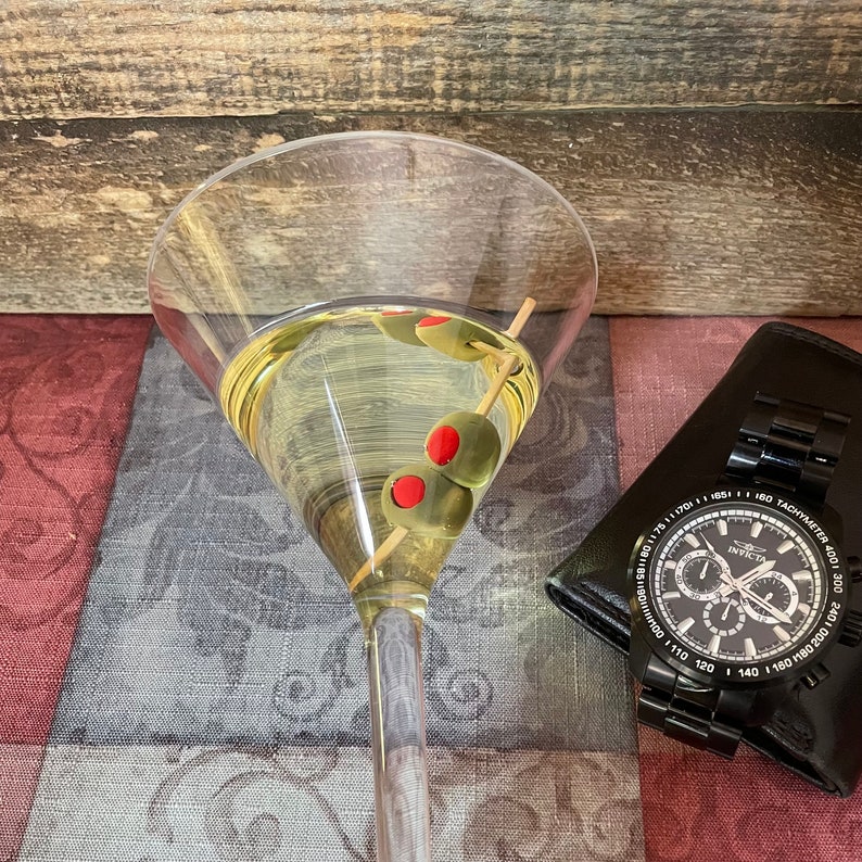 Op de afbeelding: Een martini-glas gevuld met een heldere vloeistof en groene olijven. De olijven hebben rode centra. Het glas staat op een gepatroneerd stofoppervlak. Een zwart horloge met een zwarte wijzerplaat en witte wijzers bevindt zich op de achtergrond.