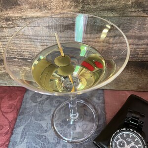 Op de afbeelding: Een martini-glas gevuld met een heldere vloeistof en twee groene olijven op een tandenstoker. Het glas staat op een gepatroneerd oppervlak met een zwart horloge op de achtergrond.