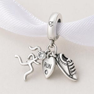May include: A silver charm featuring a running figure, a heart pendant with the word "RUN", and a running shoe. The charm is suspended from a loop with a heart design and small clear crystals. The charm is on a white ribbon.