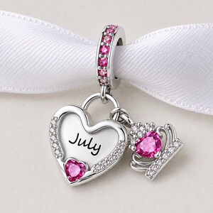 May include: A silver charm with a heart-shaped pendant that says "July" and a small crown. The heart and crown have pink gemstones. The charm is attached to a silver ring with pink gemstones.