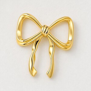 Puede incluir: Broche dorado en forma de lazo. El lazo tiene un diseño detallado con líneas curvas y un nudo en el centro. El broche es probablemente de metal y tiene una superficie brillante y reflectante. Los extremos del lazo están suavemente curvados.