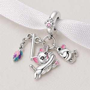 May include: A silver charm featuring a playful cat, a mouse, and a colorful feather. The cat has pink accents and is reaching out. The mouse has pink ears and a tail. The charm is attached to a silver loop with a paw print.