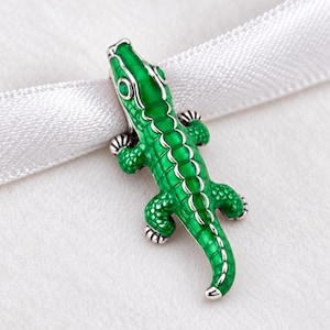 Pode incluir: Um berloque de prata em forma de crocodilo com detalhes em esmalte verde vibrante nas costas e na cabeça. O crocodilo apresenta escamas texturizadas, quatro patas e uma cauda longa, com uma pequena pedra verde cravada como olho na cabeça.