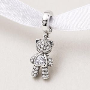 May include: A silver teddy bear charm pendant with a heart-shaped crystal on its chest. The bear is covered in small, sparkling crystals. The charm hangs from a silver loop with a small crystal and is attached to a white ribbon.