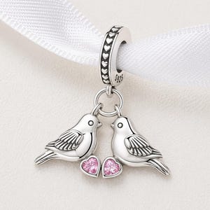 May include: A silver charm featuring two birds facing each other, with pink heart-shaped gems below. The charm has a loop adorned with heart-shaped cutouts and is attached to a white ribbon. The charm is marked with "S925".