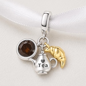 Może przedstawiać: Srebrny charms z czajniczkiem, brązowym okrągłym kamieniem i złotym rogalikiem. Na czajniczku napis "Tea". Charms zawieszony na srebrnej pętli.