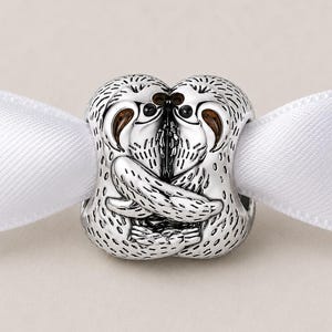 May include: A silver charm featuring two sloths embracing. The charm has detailed fur texture and brown accents on the faces. A white ribbon is threaded through the charm, suggesting it could be a pendant or a decorative element.