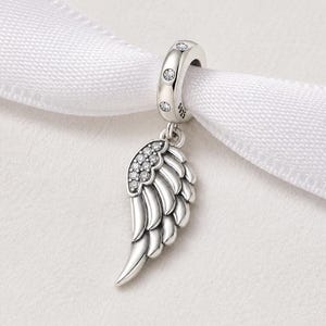 May include: A silver charm with an angel wing design, adorned with sparkling clear crystals. The charm is suspended from a ring with three small, round crystals. The wing features detailed feathering and is attached to a white ribbon.