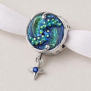 May include: A silver charm with a celestial design. The charm features a swirling pattern of blue and green glitter, accented with a dangling star pendant with a blue gemstone. The charm is attached to a white ribbon.