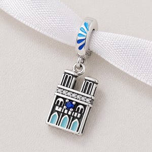May include: A silver charm depicting the Notre Dame Cathedral, with blue and white enamel detailing. The charm has a detailed design, a blue gemstone, and clear crystals. It hangs from a silver bail with blue enamel accents.