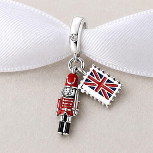 May include: A silver charm featuring a red, white, and black enamel nutcracker soldier and a rectangular postage stamp charm with a red, white, and blue Union Jack flag design, suspended from a silver bail with a small embedded clear stone.