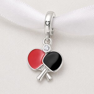 May include: A silver charm featuring two crossed ping pong paddles, one red and one black, with a small diamond accent. The charm hangs from a silver loop with a heart detail, perfect for a bracelet or necklace.