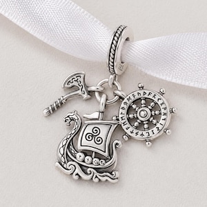 Op de afbeelding: Een zilveren bedelarmbandhanger met Viking-thema bedels. De bedels omvatten een Vikingschip, een bijl en een scheepsstuur. De hanger is opgehangen aan een wit lint.