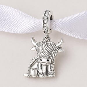 Puede incluir: Un charm de plata con un dise&ntilde;o de vaca de las Tierras Altas. El charm presenta una representaci&oacute;n detallada de la vaca con cuernos y pelo largo. El lazo superior est&aacute; incrustado con peque&ntilde;as piedras transparentes y cuelga de una cinta blanca.