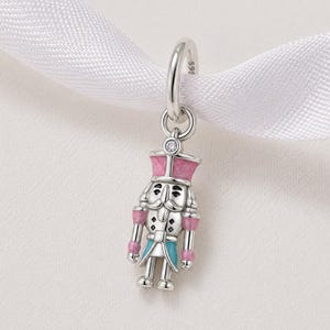 May include: A silver-coloured charm depicting a nutcracker soldier. The charm features a pink hat and accents, a light blue jacket, and silver details. A small, clear gem is set above the charm. The charm is suspended from a silver loop.