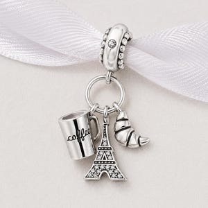 May include: A silver charm featuring a coffee cup, Eiffel Tower, and croissant suspended from a ring. The coffee cup is inscribed with the word "coffee". The Eiffel Tower is embellished with small stones. The charm hangs from a white ribbon.