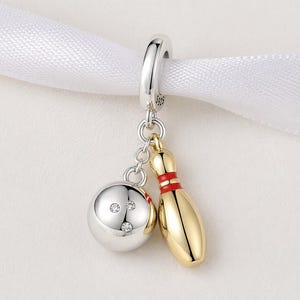 Könnte beinhalten: Ein silberner Charm-Anhänger mit einem Bowlingball mit drei klaren Steinen und einer goldfarbenen Bowlingkegel mit rotem Streifen. Der Anhänger hängt an einer silbernen Schlaufe, ideal für ein Armband oder eine Halskette.