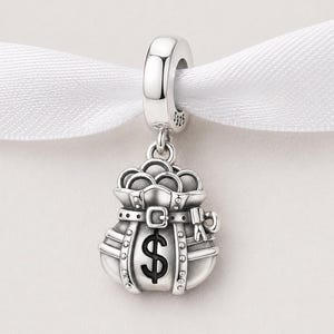 Op de afbeelding: Een zilveren bedel met een geldbuidelontwerp. De tas is gedetailleerd met een riem, klinknagels en een dollarteken. De bedel hangt aan een lus, perfect voor een armband of ketting.