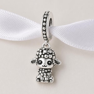 May include: A silver sheep charm with a textured body and small ears, suspended from a beaded ring. The sheep has a cartoonish design, with a smiling face and large eyes. The charm is attached to a white ribbon. The charm is made of silver.