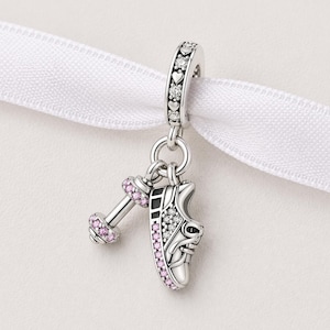 Könnte beinhalten: Silberner Charm mit einem Hantel- und einem Laufschuh-Anhänger. Die Hantel hat rosa Akzente, und der Schuh ist mit weißen und rosa Edelsteinen verziert. Der Charm ist an einem silbernen Band mit Herz- und Edelsteindetails befestigt.