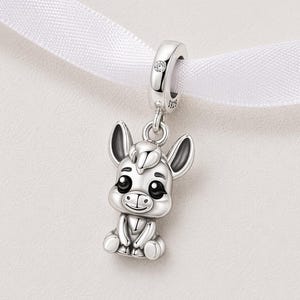 Puede incluir: Un charm de plata con un diseño de burro de dibujos animados. El charm tiene un pequeño lazo con una gema transparente y está sujeto a una cinta blanca. El burro tiene ojos grandes y está sentado.