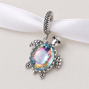 May include: A silver-toned turtle charm with a large, iridescent, multi-colored crystal shell. The charm has detailed flippers and a textured loop for attaching to a bracelet or necklace. The charm is suspended from a white ribbon.