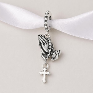 Può includere: Un charm in argento raffigurante mani in preghiera, un rosario e una croce. Il charm è sospeso a un anello di perline. Il design è dettagliato e ha una finitura lucida. Il charm è probabilmente per un braccialetto o una collana.
