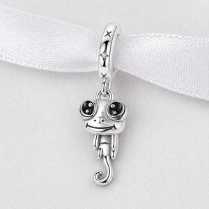 Könnte beinhalten: Ein silberner Charm-Anhänger mit einem verspielten Frosch-Design. Der Anhänger zeigt einen Frosch mit großen, schwarzäugigen Augen, einem lächelnden Mund und einem eingerollten Schwanz. Der Anhänger hängt an einer silbernen Schlaufe mit Sternakzenten, perfekt für ein Armband oder eine Halskette.
