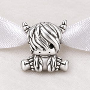 May include: A silver charm bead shaped like a Highland cow with long, textured hair, two small horns, and a smooth finish. The charm features a central hole for threading onto a bracelet or necklace, showcasing detailed metalwork and a polished surface.