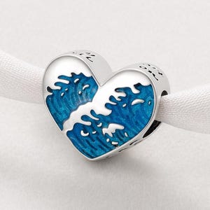 May include: A silver-coloured heart-shaped charm featuring a blue and white wave design. The charm is designed to be threaded onto a bracelet or necklace. The wave design is detailed and striking.