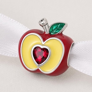 May include: A red apple charm with a yellow center and a heart-shaped red gem. The charm has a green leaf and a silver stem. The apple is on a white ribbon.