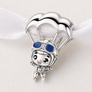 May include: A silver charm featuring a cartoon parachutist. The figure wears a helmet with blue goggles and a jumpsuit. The parachute is silver with a cloud design. The charm is attached to a white ribbon.